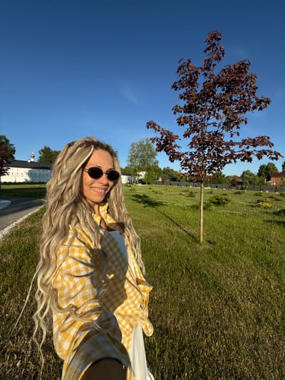 An adult female is captured in a selfie-style outdoor photograph in Dumino, Russia, under clear blue skies with bright sunlight. She is positioned in the foreground, smiling with teeth visible, and wears round dark sunglasses. Her long, wavy blonde hair is styled with numerous dreadlocks or braided extensions. She is dressed in an unbuttoned yellow and white gingham long-sleeved shirt over a white top. To her left, a light-colored paved pathway curves through a well-maintained grassy area. In the background on the left, a large, white building with a distinctive architectural style, including golden domes or cupolas on its roof, is visible, possibly a monastery or church, with an extending white wall. To the right, a young maple tree with reddish-brown leaves stands in a field of green grass dotted with smaller shrubs and ornamental plants. Further in the background, a dark fence line separates the manicured area from scattered residential-style buildings and additional trees, indicative of a village or suburban environment.