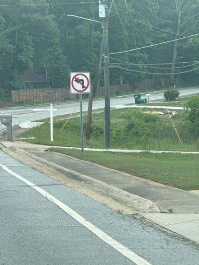 The image shows a "no left turn" traffic sign next to a road in Mountain Park, United States of America. The sign is red and white, mounted on a metal pole. A concrete curb is visible on the right side of the road, and green grass and trees are in the background. Power lines are strung overhead. A white cross is planted in the grass.
