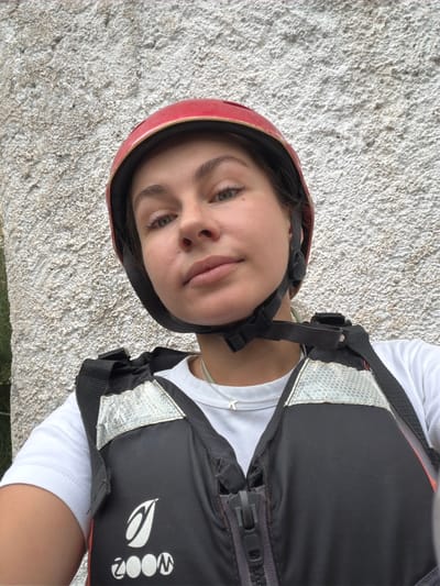 A young woman, wearing a red helmet and a black life vest over a white t-shirt, poses for a selfie in front of a textured, light-colored wall. The life vest has reflective silver detailing and a prominent white logo with the text "ZOOM". The woman has light brown hair, green eyes, and is looking directly at the camera. The background appears to be a natural rock formation or an unplastered concrete surface, possibly in an outdoor setting. The image is taken from a slightly low angle, looking up at the woman.