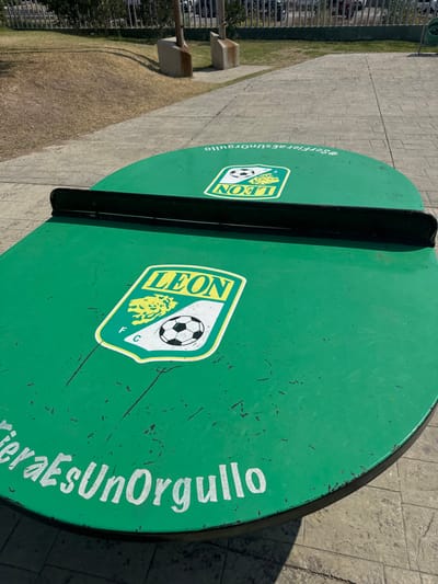 This image depicts an outdoor, green, round-ended table tennis table located in La Venta, Mexico. A black net spans the table's width. The table's surface shows multiple scratches and scuff marks. Two identical logos are prominently displayed on the green surface: each features "LEON" in yellow capital letters, a yellow silhouette of a lion's head, and a black and white soccer ball, all enclosed within a white shield with a green border. The letters "FC" are also visible within the logo. White text along the curved edge of the table reads "#SerFieraEsUnOrgullo". The immediate ground surrounding the table is a grey, textured paved surface, partially covered by the table's shadow. In the background, beyond the paved area, lies a patch of dry, brownish-yellow grass. A dark-colored metal fence separates this grassy area from what appears to be a parking lot or street, where several light-colored vehicles are discernible. Two concrete support structures are visible in the grassy area. The scene is illuminated by bright sunlight, casting distinct shadows. No individuals are present, and no actions are being performed.