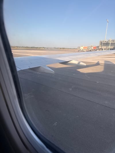 The image shows an airplane wing from the perspective of a passenger inside the cabin, looking out the window. The plane is parked on the tarmac at Alicante-Elche Airport (ALC) in Elx / Elche, Spain. The wing, which is white, extends across the frame from the left. Beneath the wing is a dark gray asphalt surface, typical of an airport runway or taxiway. In the background, several airport buildings and jet bridges are visible. The sky is clear and blue, indicating good weather conditions. The lighting suggests it is either early morning or late afternoon due to the long shadows cast on the tarmac by the wing. There are no people clearly visible in the foreground, though a few figures might be seen near the jet bridges in the distance. The overall scene depicts a typical airport environment during boarding or deplaning.