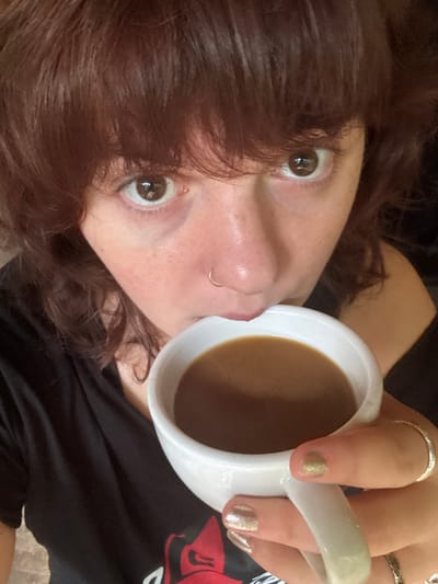A female subject is depicted in a close-up, high-angle portrait, located in Flores, Guatemala. The subject has reddish-brown hair with bangs, brown eyes looking directly at the camera, and fair skin, with a silver nose ring in the left nostril. She holds a white ceramic mug to her lips, containing a light brown beverage. Her right hand, featuring nails with gold glitter polish and two silver rings on the ring finger, grips the mug. She wears a black short-sleeved top with a red and white graphic partially visible on the chest. The background is dark and out of focus.