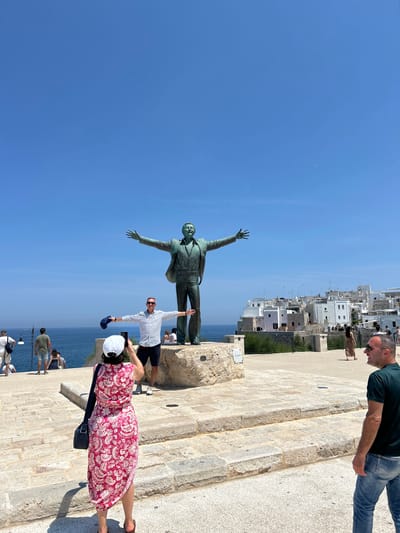 The image depicts a statue of Domenico Modugno, the singer of "Volare," located in Polignano a Mare, Italy. The statue, made of bronze, stands on a stone pedestal overlooking the Adriatic Sea. In the foreground, a man in a striped shirt and shorts poses with his arms outstretched, mimicking the statue's stance. To his left, a woman in a pink floral dress is taking a photograph of him with her phone. Behind them, other tourists are visible in the background, enjoying the view of the sea and the white buildings of the town. The sky is a clear blue, and the sun is shining brightly. The overall scene is one of leisure and tourism, with people enjoying the iconic landmark and the beautiful coastal scenery.