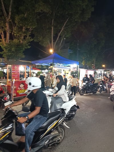 This media file captures a bustling night scene at a food market in Bandarbireuen, Indonesia. The primary subjects are people, many of whom are riding or near motorcycles. Several food stalls are visible, adorned with colorful signs and under blue and red tents, suggesting a vibrant commercial atmosphere.
The activity taking place is a food market, with people browsing and purchasing food. The presence of numerous motorcycles indicates that many visitors have arrived by this mode of transport, and the overall impression is one of lively commerce and social gathering. The time of day is clearly night, illuminated by artificial lights from the stalls and street lamps, casting a warm glow on the scene. The weather appears to be clear, as evidenced by the unhindered visibility.
Notable details include a food stall with a sign featuring a cartoon character and text that appears to be in Indonesian, possibly advertising dim sum. People are dressed in casual attire, with some women wearing hijabs, which is common in Indonesia. The emotion conveyed is one of energetic activity and community. The ground is paved, suggesting a street or designated market area.
The presence of motorcycles, the types of food stalls, and the clothing of the people are strong indicators of the Indonesian setting. The overall mood is lively and engaging, characteristic of a popular local market.
