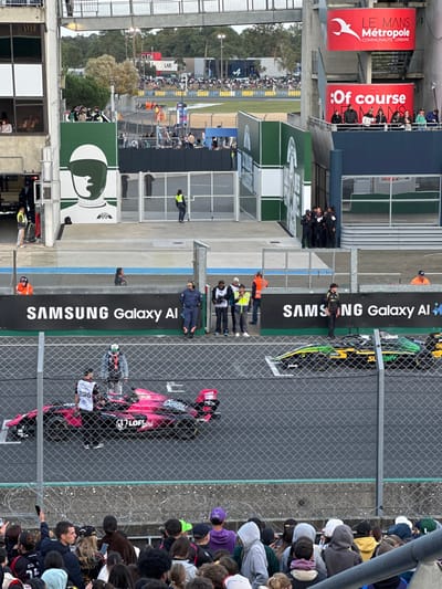 The image captures a motor racing scene at the Circuit des 24 Heures du Mans, located in Le Mans, France, specifically indicated by "Le Mans Métropole" signage. The perspective is from an elevated spectator area overlooking the track.
In the immediate foreground, a dense crowd of spectators, predominantly young adults, are visible from behind, standing and observing the track. They are separated from the circuit by a chain-link fence with grey vertical posts and a low concrete barrier.
On the track level, two open-wheel race cars are positioned. The prominent car on the left is pink/magenta with "Gentle," "LOF girl," and "Skatte" branding. A person in a white top and black shorts stands beside its rear wing. To the right, a second open-wheel car, green and black with yellow accents, is partially visible. Behind the track, a dark grey barrier displays "SAMSUNG Galaxy AI" branding. Several trackside personnel are positioned behind this barrier; some wear orange safety vests, one is in blue overalls, and others wear dark uniforms, observing the scene.
Further back, on the left, a large building section features a green panel with a white graphic of a helmeted driver's profile. Above this, a multi-story building with windows houses more spectators. A metal pedestrian bridge spans across the track area. On the right, a blue building structure with multiple windows and balconies also hosts spectators. Attached to this structure are red banners displaying "LE MANS MÉTROPOLE COMMUNAUTÉ URBAINE" and "Of course."
In the background, beyond the immediate track structures, a dense line of trees is visible. A white container marked "LAB" and "ALPINE" signage are discernible, along with distant crowds and vehicles, suggesting other parts of the circuit. The sky is overcast.