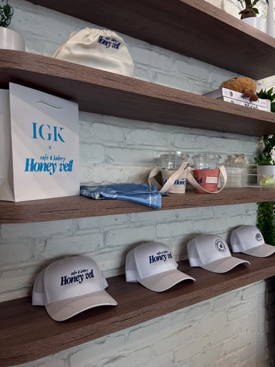 The image shows shelves in a shop displaying merchandise. On the top shelf, there is a white drawstring bag with "Honey veil cafe & bakery" printed in blue. To the right, a stack of books with a small stuffed animal on top.
The second shelf features a white paper shopping bag with blue text reading "IGK x cafe & bakery Honey veil". Next to it are two clear plastic cups with lids, each inside a fabric sleeve. One sleeve is beige and the other is coral. A folded blue cloth item is also on this shelf.
The bottom shelf displays several white mesh-backed baseball caps with blue embroidery. The caps have logos and text that appear to be related to "Honey veil cafe & bakery".
The background is a white brick wall. The setting is likely a retail store, possibly a cafe or boutique, located in South Miami, United States of America.
