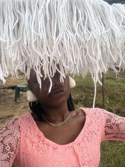An outdoor portrait features a person, likely a woman, with dark skin, wearing a pink dress adorned with intricate lace patterns. A prominent gold chain necklace is visible around her neck. Her hair or headwear is characterized by numerous thick, white, yarn-like strands that frame and partially obscure her face. Her eyes appear to be closed or looking downward, and she has a nose piercing. A small, white decorative item, possibly a flower, is tucked near her shoulder.

The background shows natural elements, including green grass and patches of dirt, suggesting an outdoor location. A small green plastic chair is visible further back in the scene. The lighting indicates daylight conditions. The person seems to be posing for the photograph in a casual setting. No text is visible in the image. The location is Aghalokpe, Nigeria.