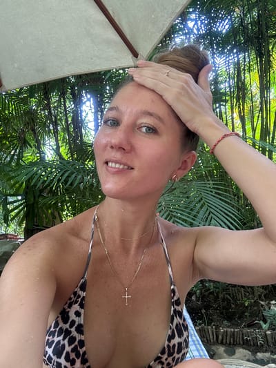 A woman with her hair in a bun poses for a selfie outdoors, beneath a large umbrella. She is wearing a leopard-print bikini top, a gold necklace with a cross pendant, and a bracelet. Her left arm is raised, resting on her head. Her skin is wet with water droplets. The background consists of lush green foliage and bamboo stalks, illuminated by sunlight filtering through the leaves. The setting appears to be a tropical environment, potentially near Khlong Thom Nuea, Thailand, given the vegetation.