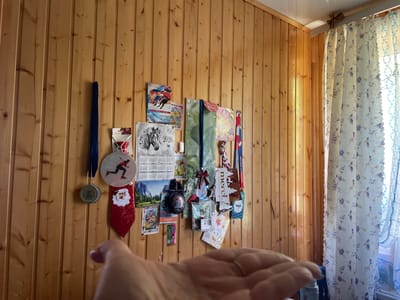 The image shows a wall made of light-colored wood planks, possibly in a room in Первомайский, Russia. Various objects are displayed on the wall, suggesting a collection of memorabilia.
Prominent items include:
- A blue lanyard with a medal hanging from it, the medal has text that appears to be in Russian and says "ВСЕ НА ЛЕДІ" (Everyone on the ice).
- A silver medal with an inscription.
- A red sequined Santa Claus decoration.
- A circular ornament with a figure skating design on it.
- A calendar for the year 2026 featuring a black and white image of a horse.
- Several other small pictures, cards, and ribbons, some with text like "merci" and "ТРЕНЕР" (Coach). One ribbon has the words "ЧЕМПИОНАТ РОССИИ" (Russian Championship) visible.
- A green decorative bag with a bow.
- A blue curtain with a floral pattern is visible on the right side of the image, letting in natural light.
The overall impression is a personal display of achievements, possibly related to figure skating or winter sports, given the ice-related texts and imagery. The lighting suggests it's daytime, with light coming from the window. The focus is on the wall display, with a hand obscuring the bottom part of the image.