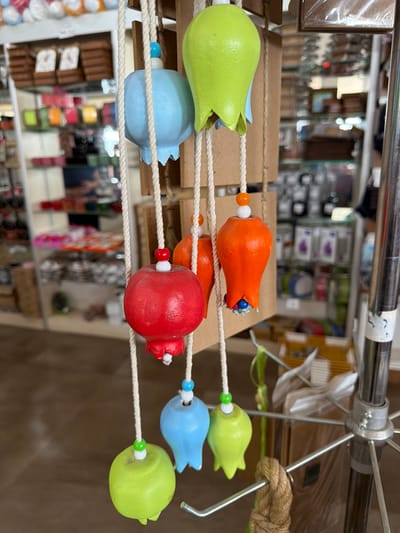 The image captures a display of colorful, bell-shaped ornaments hanging from strings. The ornaments are primarily red, orange, light blue, and lime green, and each is adorned with small beads. They are suspended in front of a store interior, which appears to be a shop selling various goods, including boxed items and shelves stocked with colorful products. The background is blurred, suggesting a shallow depth of field, and showcases racks and aisles of merchandise. The overall scene depicts a retail environment with decorative items for sale. The location is identified as Acıpayam, Turkey.