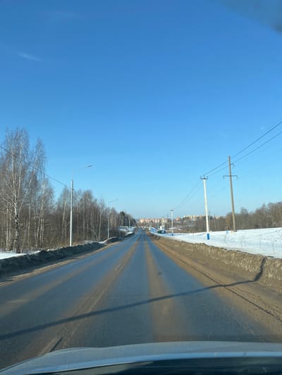 An image captured from inside a vehicle depicts a two-lane asphalt road extending into the distance under a clear, bright blue sky in Poddubki, Russia. The road surface appears partially wet or reflective, with several parallel dark stripes, likely shadows, running along its length. On the left side of the road, a line of bare deciduous trees, predominantly birches, stands alongside a snow-covered verge mixed with dirt. Multiple streetlights with horizontal fixtures are positioned along this side. Overhead power lines traverse the scene from the left. On the right, the roadside features snow-covered ground with patches of exposed dirt and a few small, bare shrubs. Several utility poles, one white and one brown, support multiple power lines running parallel to the road. Further along the right, blue-colored structures are visible next to the road. In the mid-to-far distance, a cluster of multi-story residential buildings is visible on a slight elevation, flanked by forested areas. The immediate foreground shows the upper edge of a vehicle's dashboard and windshield. The environment suggests a winter setting with sunny weather. No people are visible.