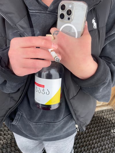 An individual is depicted from the upper chest to mid-thigh, wearing a dark grey hooded sweatshirt, a black zippered puffer vest with a small white polo horse logo on the right chest, and light grey sweatpants. The person's right hand holds a lit cigarette, with a visible ash tip and rising smoke, between the thumb and index finger. A small, dark, swirling tattoo is present on the back of the right hand. The left hand holds an iPhone, visible in a clear case with a MagSafe ring. Tucked against the person's torso, secured by the left arm, is a dark bottle with a white and yellow label. The label features Georgian text: "ჭაჭა," "ტკბილი," "ალკოჰოლი 45%," and "0.5ლ." The individual stands on a dark, textured mat. The scene is located in Juta, Georgia.