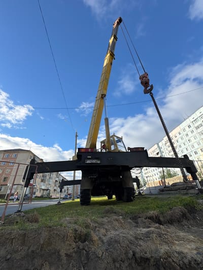 The image depicts an outdoor scene, possibly during the daytime, under a bright blue sky with scattered white clouds. The main object in the foreground is a yellow crane with its boom extended upwards and to the left. A metal chain with a hook attachment is visible at the end of the boom. The crane is mounted on a heavy-duty truck, part of which is visible, including its tires and chassis.

In the background, there are multi-story residential buildings, characteristic of a city or town. The buildings appear to be made of concrete and have many windows. A road is visible between the crane and the buildings, with some parked cars. The ground in the immediate foreground appears to be disturbed earth or a trench, with grass growing along its edge.

A license plate with Cyrillic characters and numbers is visible on the back of the truck, reading "P 745 CX 11". The overall impression is of a construction or maintenance activity taking place in an urban or suburban environment. There are no people clearly visible in the image, though a small, distant figure in pink might be a person. The weather appears to be fair and sunny. The context provided indicates the location is Usinsk, Russia.