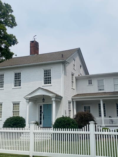 A white, multi-story residence stands behind a white picket fence in Ridgefield, United States. The house features white clapboard siding and a dark shingled roof. A red brick chimney topped with a black weathervane depicting a horse and rider is visible on the main section of the roof. The primary entrance has a light blue door under a portico supported by two white columns, with a black lantern hanging above. Numerous white-framed, multi-paned windows are present across both stories of the main structure and an attached wing to the right. The attached wing also features a covered porch with white columns. Green, rounded bushes are positioned near the front entrance and a darker, spherical bush is in front of the attached wing. Green grass is visible between the fence and the house. A large green tree is partially visible on the far left. The sky is light grey and overcast. No individuals are present.