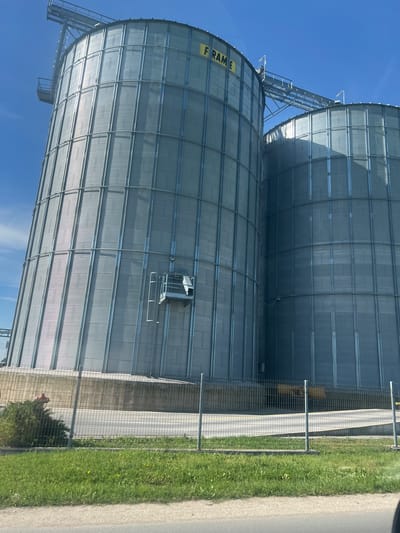 Two large metal grain silos are visible against a blue sky. The silos are cylindrical with corrugated metal siding and a network of metal supports. One silo has the word "FRAME" printed vertically on its side. A metal walkway and ladder system connect the silos. The silos are bordered by a chain-link fence, a concrete or paved area, and a grassy strip with some wildflowers. Beyond the grass is a section of pavement. The scene appears to be in an agricultural or industrial area, potentially in the city of Brezovo, Bulgaria. There are no people visible.