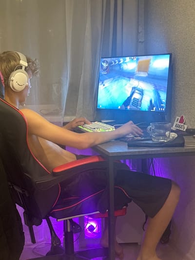 In the city of Sredneuralsk, Russia, a young person is captured in a dimly lit room, immersed in a gaming session. The individual, seen from the side and slightly from the back, is seated in a black and red gaming chair, wearing white headphones with a smiley face design. They are looking intently at a computer monitor displaying a video game, their right hand positioned over a white keyboard. The room is illuminated by purple ambient lighting emanating from beneath the desk and from a PC case with a fan visible at the bottom right. On the desk, next to the monitor, sits a clear glass cup with some liquid and ice cubes, and a small sticker with Russian text and a heart icon. The background features a sheer curtain and a textured wallpapered wall.