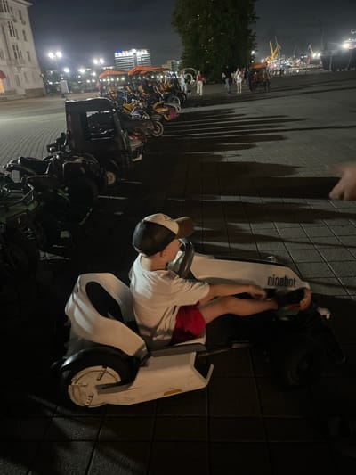 An elevated view shows a boy wearing a white t-shirt, red shorts, and a black and white baseball cap, sitting in a white and black electric go-kart with the brand name "ninebot" visible on its side. The boy's hands are on the steering wheel. The go-kart is on a dark tiled pavement, casting shadows from streetlights. In the background, several other electric scooters and small vehicles are parked, some with canopies. Beyond the vehicles, a wide waterfront promenade with people walking and a port with cranes and ships can be seen under a dark night sky. The location is likely a public recreational area in a city, possibly Berezhnoi, Russia, given the presence of a port.