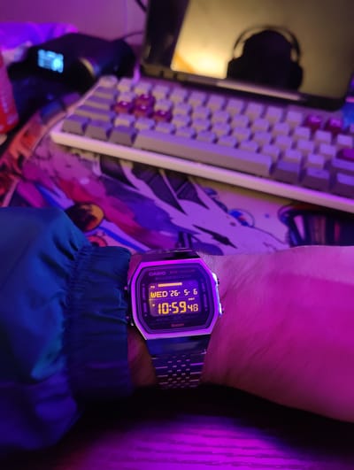 A photograph taken indoors under purple lighting shows a person's wrist with a digital Casio watch displaying the time 10:59:48 on Wednesday, May 26th. The watch band is made of metal. The sleeve of a blue jacket or shirt is visible to the left of the watch. In the background, a keyboard with white keys and red accent keys sits on a multicolored mousepad. A monitor and headphones are visible behind the keyboard, but the location in Dorst, Netherlands cannot be determined from the image.