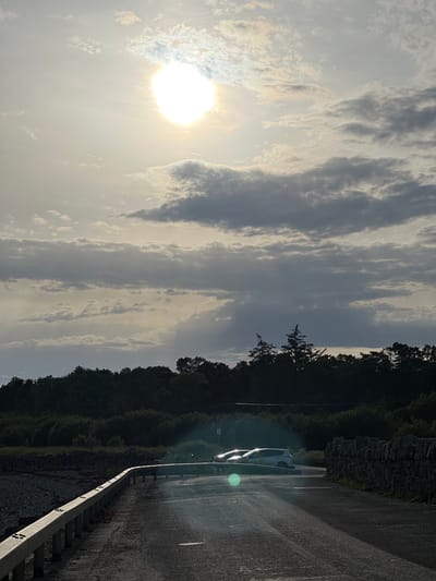This is a photograph taken in Terhillion, Ireland, likely during the late afternoon or early evening due to the low angle of the sun and the long shadows. The weather appears to be partly cloudy, with the sun breaking through scattered clouds that create a streaky, dramatic effect in the sky.

The scene depicts a roadside setting. On the left side of the image, a dense line of trees forms a dark backdrop against the bright sky. A rough stone wall runs along the edge of the road, adding a rustic element to the landscape.

The road itself is paved and appears slightly damp, possibly from recent rain, and reflects the light from the sun and any streetlights that might be present but are not clearly visible. A silver car is parked on the side of the road, its headlights on. The car is positioned on the right side of the frame, facing away from the viewer and towards the distance. A metal guardrail with reflective markers lines the edge of the road, separating it from the embankment.

There are no people visible in the image. The overall mood is serene and contemplative, with the natural beauty of the landscape enhanced by the atmospheric lighting. A small, faded sign is visible on a post near the parked car, but the text is illegible.