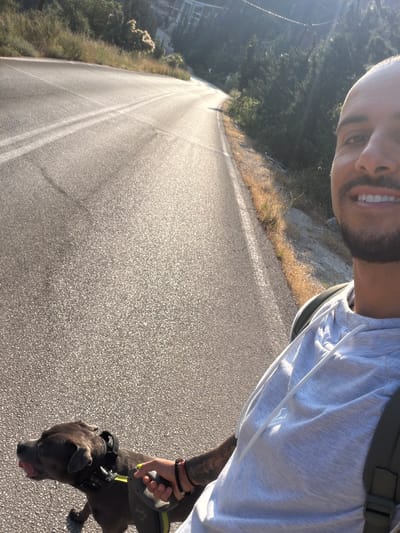 A selfie shows a man and a dog walking on a road. The man, partially visible in the upper right, has a shaved head, a light gray t-shirt, and a dark-colored backpack. He has a light beard and is smiling. His left arm is visible, with a tattoo and beaded bracelets on his wrist, holding a black and yellow retractable leash. The dog, a dark gray breed, is walking to the right with its tongue slightly out. The road is gray asphalt with white lane markings. To the left of the road, there is green vegetation and trees. The location is near the city of Municipal Unit of Sfakiates, Greece.