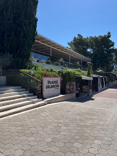 The image depicts the exterior of a restaurant named "[PLACES] DALMACIJA by VALAMAR" in Makarska, Croatia. The restaurant features a modern architectural design with a large glass facade and an expansive covered outdoor seating area. In front of the restaurant, there are stone steps leading up to the entrance. A prominent white sign with the restaurant's name is positioned to the left of the steps. Several dining tables and chairs are visible through the glass and under the covered patio. Greenery, including trees and flowering bushes, surrounds the building. A paved walkway with hexagonal stones extends from the foreground. There are no visible people in the immediate foreground or interacting with the restaurant. The scene is set against a clear blue sky on a sunny day.