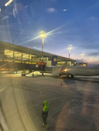 The image displays an airport tarmac scene at Krasnoyarsk International Airport, located near Емельяново, Russia, observed during either dawn or dusk. The sky overhead is dark blue, transitioning to lighter blue and subtle orange/pink hues near the horizon. Multiple tall floodlights with yellow-orange illumination cast distinct lens flares across the frame.
A white commercial aircraft, bearing "UTair" livery and the registration number "RA-73145" on its tail, is parked at a gate and connected to a dark-colored jet bridge. Several ground support vehicles are partially visible near the aircraft's left wing and the jet bridge.
One individual, clad in a high-visibility neon yellow-green hooded jacket over dark trousers, stands on the concrete tarmac in the foreground, facing away from the viewer. A second individual is discernible near the base of the jet bridge. The tarmac surface shows tire marks and features a single orange traffic cone.
In the background, the modern terminal building with a multi-story glass facade is visible, illuminated from within. A prominent sign reading "KRASNOYARSK" is mounted on the roofline of the terminal. Reflections are present on the left side of the image, suggesting observation through a window.