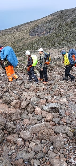 Four individuals are depicted trekking across a rocky, inclined path at Gilman's Point, Tanzania, under an overcast sky. The terrain consists primarily of dark gray, reddish-brown, and black volcanic rock and scree, with minimal sparse vegetation.
From left to right:
1. A person partially visible, wearing orange trousers and a bright blue poncho or backpack cover over a backpack.
2. A person wearing dark attire, a white helmet or hard hat, and carrying a backpack covered by a bright yellow/lime green rain cover.
3. A person wearing a dark top, dark trousers, a white baseball cap, and carrying a dark-colored backpack with visible orange components.
4. A person partially visible, wearing a yellow jacket and carrying a backpack partially covered by blue fabric.
All individuals are walking uphill, equipped with hiking gear, suggesting an ascent or traverse in a high-altitude, barren mountain environment. The background shows a steep mountain slope with sparse vegetation and exposed rock.