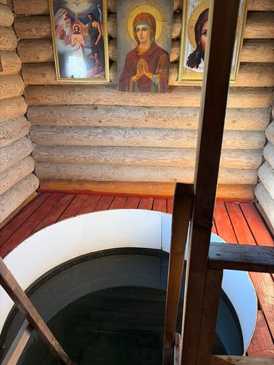 The image shows an interior view of a wooden well structure in Pobedinka, Russia. Three religious icons are mounted on the log walls. The leftmost icon depicts the Baptism of Jesus, with angels and John the Baptist present. The central icon is of the Virgin Mary with her hands clasped in prayer. The rightmost icon shows a portion of Jesus' face with a halo. Below the icons, a wooden floor transitions to a red-painted surface surrounding the opening of a well. A white curved barrier lines the well opening. A wooden staircase descends into the well, which appears to contain dark water. The walls are constructed from rough-hewn logs.