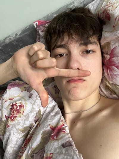 The image depicts a young individual, appearing to be male, positioned in a supine or reclining posture, looking directly at the camera. The person has short, dark brown hair and brown eyes. A thin, silver-colored chain is visible around their neck, resting on their bare upper chest. The left hand is raised, forming an "L" shape with the thumb and index finger extended; the index finger is positioned horizontally across the upper lip area. The facial expression is neutral, with slightly downturned lips. The individual is partially covered by bedding featuring a floral pattern with white, red, pink, purple, and green motifs. A solid, dark grey surface, possibly a pillow or headboard, is visible behind the person's head on the left side of the frame. The setting is indoors, consistent with a residential bedroom. The precise location within the city of Zainsk, Russia, cannot be determined from the visual information provided.