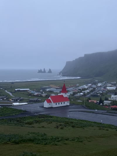 The image depicts Vik i Myrdal, a village in Iceland. In the foreground is Vikurkirkja Church, identifiable by its red roof and white walls. The church is centrally located within the town, surrounded by residential buildings. A road runs adjacent to the church. Beyond the town, the ocean stretches into the distance, partially obscured by mist or fog. Prominent rock formations rise from the sea near the shoreline. A large, steep cliff forms the backdrop, also partially obscured by fog. Green vegetation covers the areas surrounding the town. The image captures a landscape typical of coastal Iceland, characterized by rugged cliffs, coastal towns, and variable weather.
