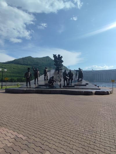 A statue depicting several figures stands on a paved area, with a dam and a large body of water in the background. The figures in the statue appear to be engaged in some sort of work or discussion. One figure, in the foreground and to the left, stands with his arm raised. Next to him, another figure is standing with his arm extended upwards, possibly pointing or gesturing. Further to the right, a figure is bent over, seemingly examining something on the ground. Another group of figures stands to the right, with one appearing to operate a surveying instrument, and another holding a rolled-up document. Behind the statue, a massive dam stretches across the water, with a hydroelectric power plant visible on its surface. The scene is set against a backdrop of lush green mountains and a clear blue sky with scattered white clouds. The sun is shining brightly, indicating it is daytime. The overall mood is one of historical significance and industrial achievement. The paving stones are laid in a brick pattern. There is no visible text in the image.