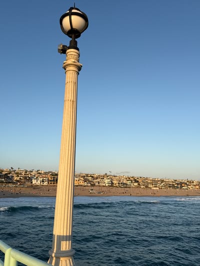 A lamppost stands prominently in the foreground of this vertical, medium-shot image. The lamppost is made of a light-colored, fluted material and is topped with a white, spherical light fixture encased in black metal. To the left of the light fixture, a small, square electrical box is attached to the lamppost. The lamppost casts a distinct shadow on the right side of its shaft.
The background reveals a wide, sandy beach populated by numerous people engaged in various activities, such as sunbathing and strolling. A lifeguard tower is visible near the center of the beach. The ocean in front of the beach exhibits moderate waves with whitecaps. Beyond the beach, a dense collection of buildings, primarily houses and multi-story residences, forms a backdrop that slopes upwards towards a clear, light-blue sky. The overall scene suggests a pleasant day at a coastal city. Based on the architecture of the buildings and the presence of palm trees, this location is likely Manhattan Beach, California, United States of America.