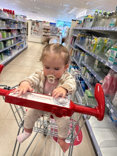 A young child, appearing to be between 12 and 24 months old, sits in a shopping cart in a Rossmann drugstore. The child has light brown hair styled in two pigtails and is wearing a light brown textured cardigan over a pink shirt and patterned leggings with pink socks. A pacifier is in the child's mouth. The child's hands rest on the red handle of the shopping cart, which displays the "ROSSMANN Mein Drogeriemarkt" logo. The shopping cart is positioned in an aisle stocked with various household and personal care products on both sides. The flooring appears to be tiled. The store is brightly lit with fluorescent ceiling lights. The location is identified as a Rossmann drugstore in Braunfels, Germany.