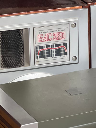 The image shows a close-up of a vintage audio component, likely a speaker or amplifier, with Cyrillic text and a frequency response graph. The component is silver with a dark brown top. The text reads "СИСТЕМА АКУСТИЧЕСКАЯ" (Acoustic System) and "БАС 220" (Bass 220). A graph displays a red line indicating frequency response across the range of 50 Hz to 20000 Hz. A circular grill with a honeycomb pattern is visible to the left of the label. The item appears to be part of a larger electronic system. The background is out of focus, suggesting an indoor setting. The location of Буран Снт, Russia, is not determinable from the visual information provided.