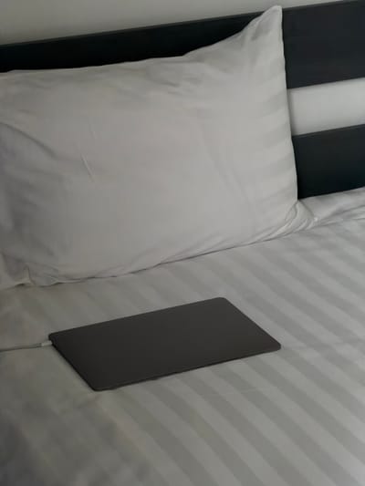 A laptop is plugged in and resting on a striped, white bed. There is a pillow behind it to its left. To the left of the pillow is a black and white headboard. The scene is indoors. There is no indication of this photo being taken in Chkalov, Armenia. No people are present.