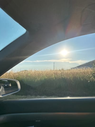 The image shows a view from inside a vehicle, looking outwards towards a sunlit rural landscape. The sun is high in the sky, casting bright rays and creating a lens flare effect. To the left, a portion of the vehicle's interior, likely the dashboard and windshield pillar, is visible, with a subtle rainbow-like refraction on its surface. To the right, the side of the vehicle and a side mirror are visible. The mirror reflects a blurred image of the interior.
The landscape outside features a field of tall, dry grass or reeds stretching towards the horizon, bathed in the warm light of the sun. A thin, vertical pole, possibly a utility pole, stands in the distance. The sky is a clear blue with a few wispy clouds. The overall impression is one of a sunny day, likely late morning or early afternoon, given the intensity of the sun and the angle of the light. The setting appears to be rural, consistent with the location context of Debelets, Bulgaria. There is no visible text in the image. The scene evokes a sense of travel and a peaceful, bright day.