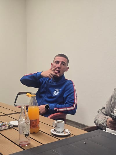 An indoor scene in Kraishte, Bulgaria, captured from a low angle, features a man striking a playful pose at a wooden table. The image is oriented sideways. The main subject, a young man with a short haircut, is wearing a blue tracksuit-style jacket with prominent white and red stripes on the sleeves and a white, possibly embroidered, emblem on the left chest. He has his tongue sticking out slightly and his left hand raised towards his face, making a gesture, revealing a ring on his index finger and a black watch on his wrist.
Seated across from him, only the arm and upper torso of another person are visible in the background, wearing a light grey jacket and holding a grey-cased smartphone.
The wooden table in front of the subjects holds several items. There's a bottle filled with an orange liquid, likely juice, with a yellow cap and a label featuring Cyrillic text. Next to it, an empty clear glass bottle with a partially visible label and a red ring at its mouth. A white ceramic cup with a distinctive black and white wavy pattern sits on a matching saucer, bearing the word "Sprema" and repeated 's' motifs. A dark, possibly black, rectangular object, possibly another part of the table or a separate item, is also visible. Partially hidden under the table items is a colorful pack, possibly cigarettes, with Cyrillic text.
The background consists of a plain, light-colored wall. The lighting appears even, suggesting an indoor setting without strong natural light. The overall mood conveyed by the main subject's expression is playful and casual.