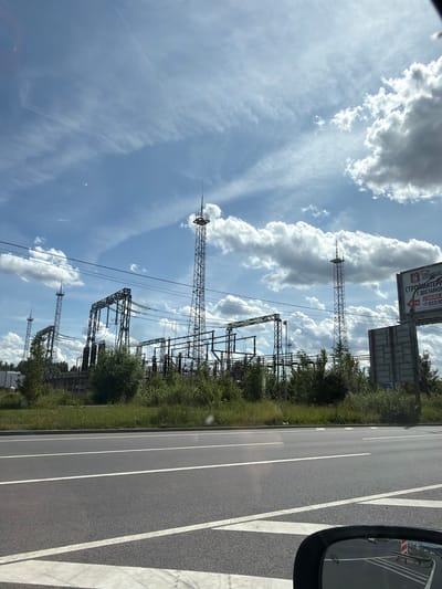 The image shows an outdoor scene with a wide asphalt road in the foreground and a power substation in the background. The sky is partly cloudy with blue patches. The power substation features several tall metal towers with electrical equipment and wires. Trees and greenery surround the substation. A billboard is visible on the right side of the image, advertising building materials. The scene appears to be located near the city of Bugry, Russia, as suggested by the presence of a billboard with Russian text. There are no people visible in the image. The perspective suggests the photo was taken from a moving vehicle.