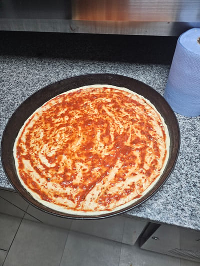 The image shows a pizza crust on a dark metal pizza pan covered with red sauce. The pan rests on a speckled gray countertop next to a roll of blue paper towels and a metal cabinet. The location is unknown, but it is suspected to be in Holywell, United Kingdom. No people are visible.