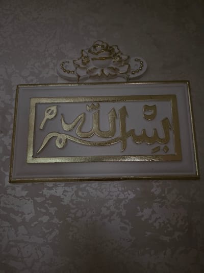 The image shows a decorative plaque with Arabic calligraphy in gold on a white background, framed by a gold border. Above the plaque, there is a white and gold ornamental piece resembling a floral design. The plaque and ornament are mounted on a textured wall with a mottled gray and white pattern. The scene appears to be an interior decoration. There is no information about the city of Shelkovskaya, Russia, present in the image.