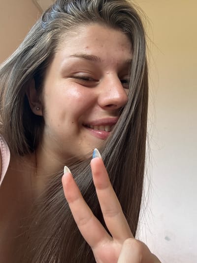 The image is a close-up, rotated photograph of a young person's face and hand. The subject has long, brown hair with lighter streaks, which falls across their face. They have fair skin with some visible blemishes and are smiling with their eyes closed. Their lips are glossy. On their right hand, they are making a peace sign, with their index and middle fingers extended. The nails are long and appear to be painted with a light, iridescent blue tip. The background is a plain, light-colored wall. The lighting suggests it is daytime, possibly indoors. The overall emotion conveyed is cheerful and playful. There is no visible text in the image.