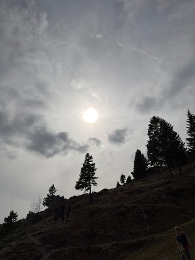 The image, taken in Kamnik, Slovenia, shows a landscape under a cloudy sky with the sun partially visible. In the foreground to the right, a steep, rocky terrain covered with vegetation is visible, punctuated by trees. Several figures, some carrying equipment, are present on the rocky surface. In the background, the sky is filled with varying cloud formations, partially obscuring the sun. Silhouetted trees are seen against the sky along the upper edge of the rocky terrain.