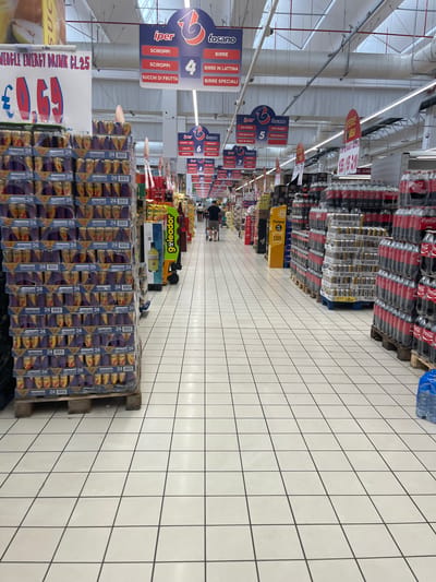 The image depicts the interior of a supermarket, likely an Iper Tosano store, given the signage. The perspective is from a low angle, looking down an aisle. On the left, stacks of canned beverages, possibly energy drinks, are prominently displayed on a pallet. These cans are labeled "24" and feature the "Eagle" branding. Further down the aisle, on the right, large stacks of bottled drinks, appearing to be Coca-Cola, are visible. Various promotional signs hang from the ceiling, advertising products and prices in Italian. One sign indicates "WEAGLE ENERGY DRINK CL 25" with a price of "€0.69". Another set of signs details product categories like "SCIROPP", "BIRRE", and "SUCCHI DI FRUTTA". People are present in the background, appearing to be shoppers, with one individual pushing a shopping cart. The floor is tiled with a white checkerboard pattern. The environment suggests a well-stocked retail space. Based on the typical locations of Iper Tosano stores, this scene could be within the city of Castiglione delle Stiviere, Italy, where an Iper Tosano supermarket is located.