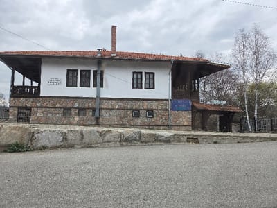 The image depicts a two-story building in the city of Nevestino, Bulgaria, viewed from a slightly elevated asphalt road in the foreground. The building features a lower level constructed with irregularly shaped, multi-colored stone blocks, and an upper level with white stucco walls. A reddish-brown tile roof covers the structure, extending outward to form covered porches or balconies on both the left and right sides of the upper floor.
On the upper floor, several dark-framed windows with multiple panes are visible. A prominent, tall brick chimney rises from the roofline on the right side of the main section. A silver-colored metal downspout runs vertically down the center of the white wall, near the chimney, discharging towards the ground level.
On the upper white wall, to the left of the downspout, a rectangular white plaque with dark lettering is affixed. Further to the right, beneath the eaves of the right-side covered area, a blue rectangular sign is visible. This sign displays white text, including "КМЕТСТВО" (Municipality/Mayoralty) and "НЕВЕСТИНО." Below this sign, a smaller, covered entrance with a red tile roof and dark wooden supports leads into the building. On the lower stone level, multiple small, horizontally oriented windows are present, along with a metal-grated window or door on the far left.
The foreground consists of a grey asphalt road and a low, rugged stone or concrete curb that separates the road from the building's immediate ground. In the background, beyond the building's right side, several bare trees and a few birch trees with white bark and early leaf growth are visible, partially obscured by a dark metal fence. The sky above is overcast with uniform grey clouds. No individuals are present in the scene.