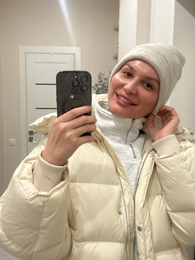 A person wearing a light-colored beanie, a gray hooded sweatshirt, and a beige puffer jacket is taking a selfie with a black smartphone. The person is smiling at the camera. The background shows a white door, a light-colored wall, and a decorative element on the wall. The location is not specified and cannot be determined from the image.