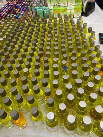 This is a high-angle, medium shot of a wall of small, clear glass bottles filled with a yellow liquid, likely perfume or cologne, each capped with a silver lid. The bottles are arranged in neat rows and columns, creating a visually striking and repetitive pattern. Some bottles have small white labels attached to them, with some text visible, including "BLUE," "ACK OPIUM," "de parfum," "Pyary," "ESCAD," "MOOD," "JAGU," "GARI," "RED," and "TILL." In the background, to the left, are stacks of green and yellow boxes, also with visible text like "Pyary" and "Best Quality," and a black electronic device, possibly a calculator or printer. The overall scene appears to be a retail or storage environment, possibly a market stall or a shop shelf, in Lamteumen Barat, Indonesia. The lighting suggests an indoor setting. There are no people visible in the frame, and the focus is entirely on the organized display of the bottled products. The sheer quantity of identical bottles suggests a commercial setting where these items are sold. The colors are predominantly yellow from the liquid, silver from the caps, and green and yellow from the boxes in the background.