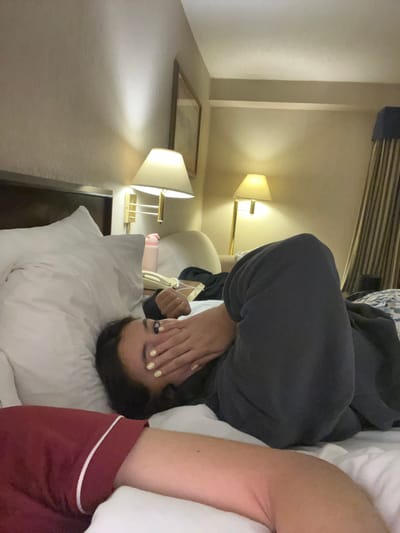 A person is lying in a bed in a hotel room. They are wearing a gray sweatshirt and red shorts with a white trim. The person's face is partially covered by their hands. The room has a lamp on a bedside table, and a picture frame hangs on the wall. A second lamp is visible in the background. The location is San Nicolás de los Garza, Mexico.