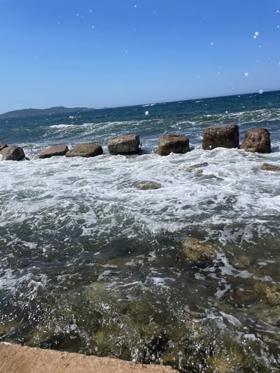 The image shows a coastline in Carqueiranne, France, under a clear blue sky. Large, rough-hewn stones, likely part of a breakwater or pier, are partially submerged in the sea. Waves are crashing against these stones, creating white foam and turbulent water. The sea is a deep blue, with a distant island or landmass visible on the horizon. The foreground features shallow water with visible rocks and a concrete or stone ledge. There are no people visible in the image.