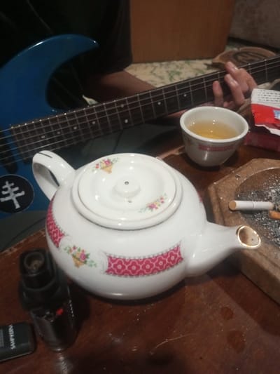 A blue electric guitar rests across a person's lap. On a wooden surface, a white teapot with floral patterns and a red design band sits beside a small cup of tea and an ashtray with cigarette butts. A lighter and a cigarette pack are also visible. The scene suggests a relaxed, perhaps creative, moment with a musical instrument and a tea break.