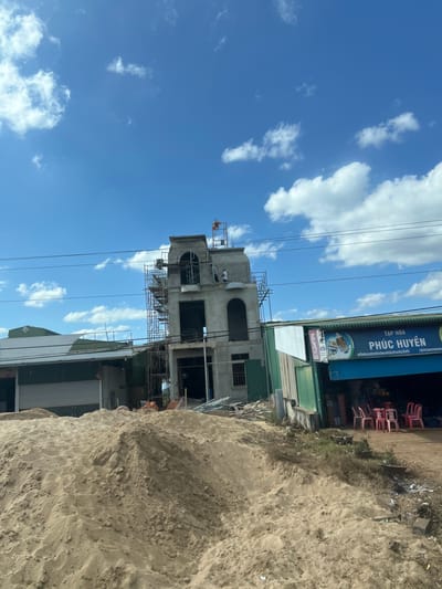 A partially constructed, two-story building stands amidst scaffolding. To the building's right is a shop with a blue sign and some red plastic chairs. A pile of dirt obscures part of the structure on the right side of the frame. Overhead, a bright blue sky is dotted with fluffy white clouds. Electrical wires run across the sky near the top of the building. The image appears to be taken from a slightly low angle, with the structures angling towards the right.